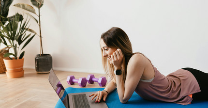 Eine Frau liegt auf einer Yogamatte und schaut lächelnd in ihren Computer. Neben ihr liegen zwei Hanteln. 