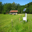 Foto: CRNS probe with solar panel installed on a partially mown meadow, with forest and a small building in the background | Foto: Cosmic Sense consortium