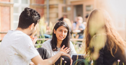 Drei Studierende sitzen auf dem Campus Neues Palais auf einer Wiese und unterhalten sich.