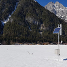 Field site Leutasch: CRNS sensor with solar panels installed at a flat valley bottom with snow. In the background are houses and forrested mountains | Photo: Cosmic Sense Consortium