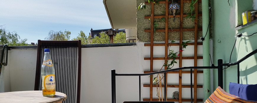 Auf einem balkon steht ein Laptio auf einem Sofa, daneben ein Tisch, ein paqar andere balkonmöbel. Es ist sonnig