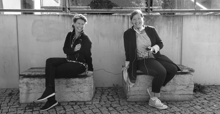 Die Produzentinnen Anne Frey und Sabine Rieder sitzen nebeneinander auf zwei Bänken. Sie halten ein Mikrofon und ein Aufnahmegerät in der Hand.