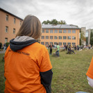 Die Helferinnen tragen orangene T-Shirt mit der Aufschrift "Kinder-Universität" und beobachten die Wiese mit den spielenden Kindern.