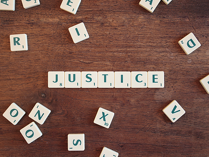 Holzfarbener Untergrund auf dem einige Scrabble Steine liegen und das Wort „Justice“ zentriert gezeigt ist.