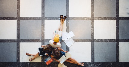 Perspektivischer Blick von einer höhren Etage in den Uniflur, wo drei Studierende auf dem Boden sitzen und auf Bücher und Laptob schauen.