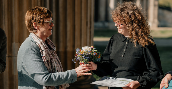 Eine Frau überreicht einer anderen eine Urkunde und einen Blumenstrauß