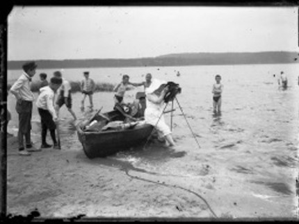 schwarz-weiß Foto einer Fotografin, die auf dem Rand eines Ruderboots sitzt und mit einer alten Kamera ein Foto machen will, dabei ist sie umringt von vielen Jungs, die teilweise in Badehosen im Wasser stehen oder bekleidet am Strand