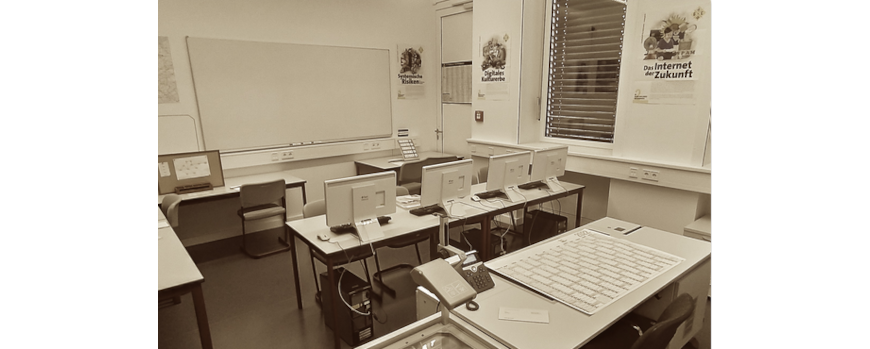 Eine Schwarzweißaufnahme von einem Seminarraum mit Schreibtischen, Laptops und einem Whiteboard.
