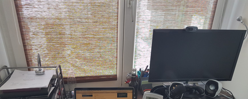Schreibtisch vor einem Fenster. in der Mitte ein aufeklappter Laptop, rechts danaben ein zweiterBildschirm und eine Tastatur, links eine Dokumentenablage
