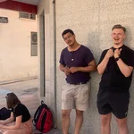 Eine Gruppe von Menschen steht, teils an einer Hauswand lehnend oder auf dem Boden sitzend, im Außenbereich. Sie wirken in ein unterhaltsames Gespräch verwickelt.