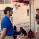 Eine Gruppe von Menschen steht, teils an einer Hauswand lehnend oder auf dem Boden sitzend, im Außenbereich. Sie wirken in ein unterhaltsames Gespräch verwickelt.