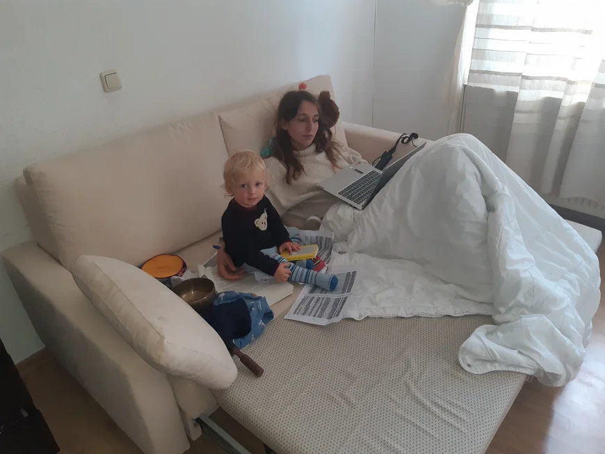 Eine junge Frau sitzt auf einem Sofa mit einer Bettdecke über den Beinen. Auf dem Schoß steht ihr Laptop. Neben ihr sitzt ein Kleinkind auf eine Haufen Ausdrucke