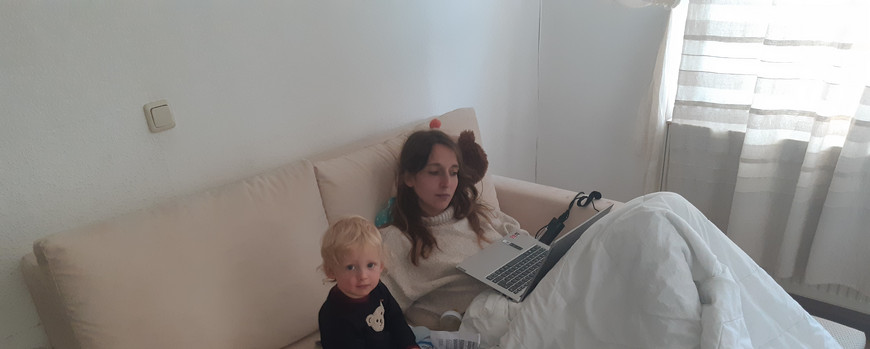 Eine junge Frau sitzt auf einem Sofa mit einer Bettdecke über den Beinen. Auf dem Schoß steht ihr Laptop. Neben ihr sitzt ein Kleinkind auf eine Haufen Ausdrucke