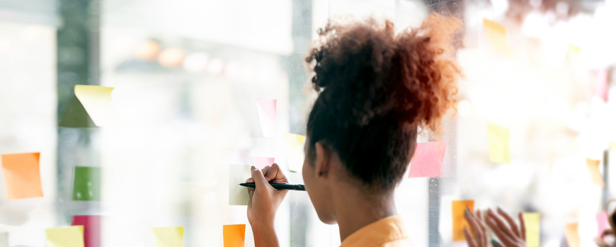 Frau vor einer transparenten Glasscheibe mit Stift in der Hand, wo Post-It Zettel dran kleben.