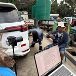 Auf dem Foto zu sehen sind im Vordergrund ein Kamerastativ, ein Hinterkopf sowie ein Laptop mit einer Textdatei. Im Hintergrund ist eine Autowerkstatt mit einer sitzenden schwarzen Person, welche interviewt wird, zu sehen.