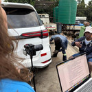 Auf dem Foto zu sehen sind im Vordergrund ein Kamerastativ, ein Hinterkopf sowie ein Laptop mit einer Textdatei. Im Hintergrund ist eine Autowerkstatt mit einer sitzenden schwarzen Person, welche interviewt wird, zu sehen.