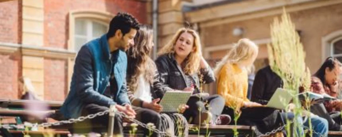 Studierende sitzen auf einer Bank am Campus Neues Palais und unterhalten sich. Eine Studentin hat einen Laptop auf dem Schoß.