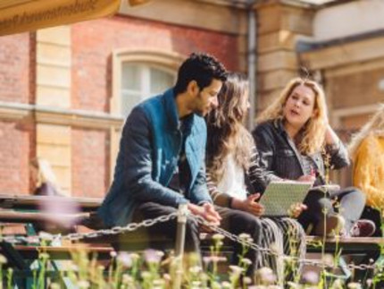Studierende sitzen auf einer Bank am Campus Neues Palais und unterhalten sich. Eine Studentin hat einen Laptop auf dem Schoß.