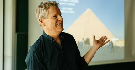 Prof. Trauth hält einen Vortrag. Er lächelt und deutet auf die Projektion von Pyramiden hinter sich.