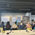 Verschiedene Menschen sitzen in einem Halbkreis vor Moderationswänden und unterhalten sich.