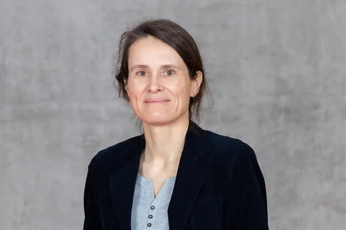 Frau mit braunen Haaren und Blazer sowie Bluse auf grauem Grund.