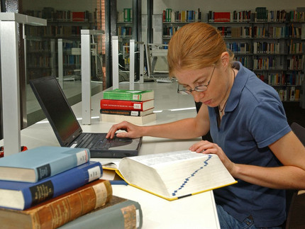Das Bild zeigt eine Frau. Sie sitzt in einer Bibliothek. Sie sitzt an einem Tisch. Auf dem Tisch steht ein Computer. Auf dem Tisch liegen sehr viele Bücher. Die Frau hat eine Hand auf dem Text im Buch. Sie will sich eine Stelle merken.