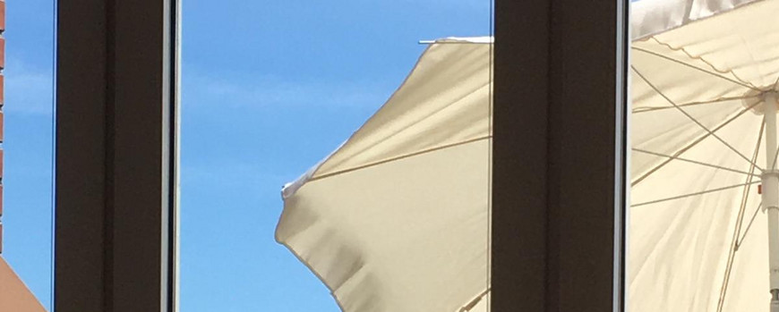 Durch eine Balkontür sieht man eine junge Frau mit ihrem Laptop auf den Knien draußen unter einem Schirm sitzen. im Hintergrund das Meer und ein Seegelboot unter blauem Himmel