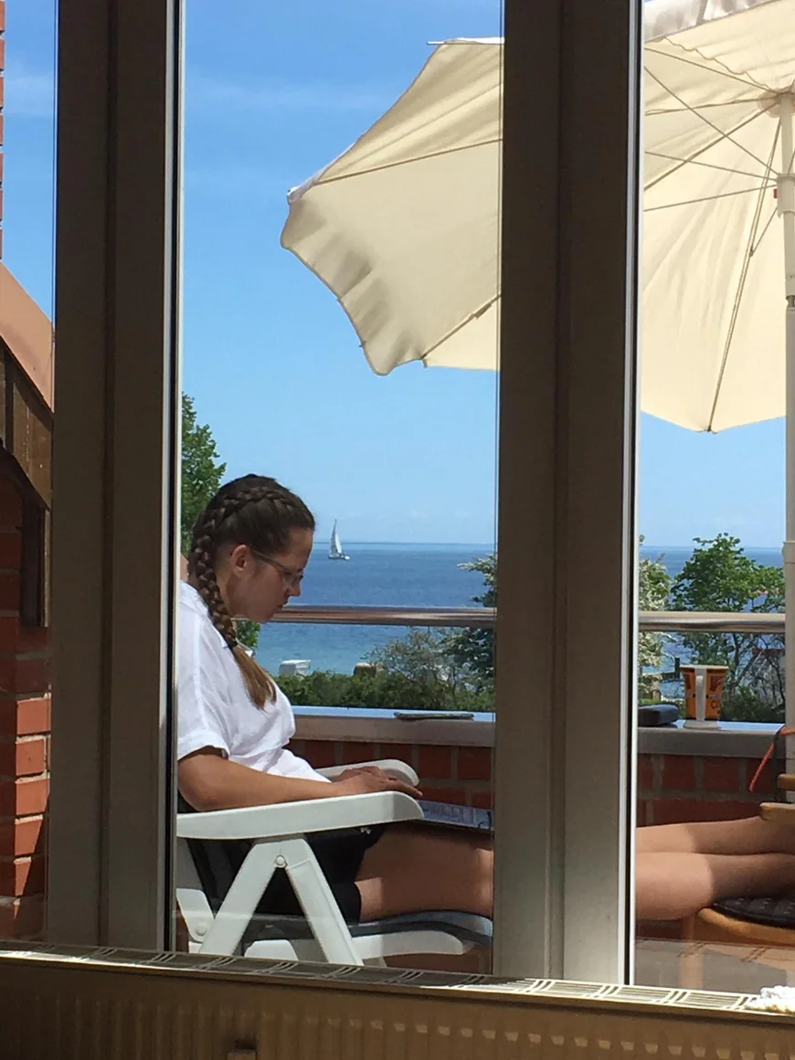 Durch eine Balkontür sieht man eine junge Frau mit ihrem Laptop auf den Knien draußen unter einem Schirm sitzen. im Hintergrund das Meer und ein Seegelboot unter blauem Himmel