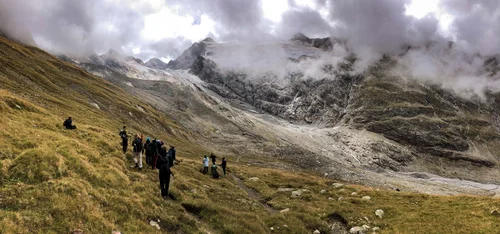 Studierende stehen auf einer Wiese, welchen auf einen steinigen Berg hinausläuft. Tief über den Studierenden hängen Wolken.