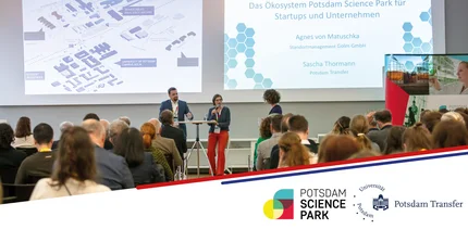 Mehrere Personen sitzen im Publikum und hören einem Mann und einer Frau, die auf der Bühne stehen, zu