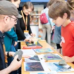 Eine Person an einem Tisch erklärt Kindern etwas zu Vulkanen