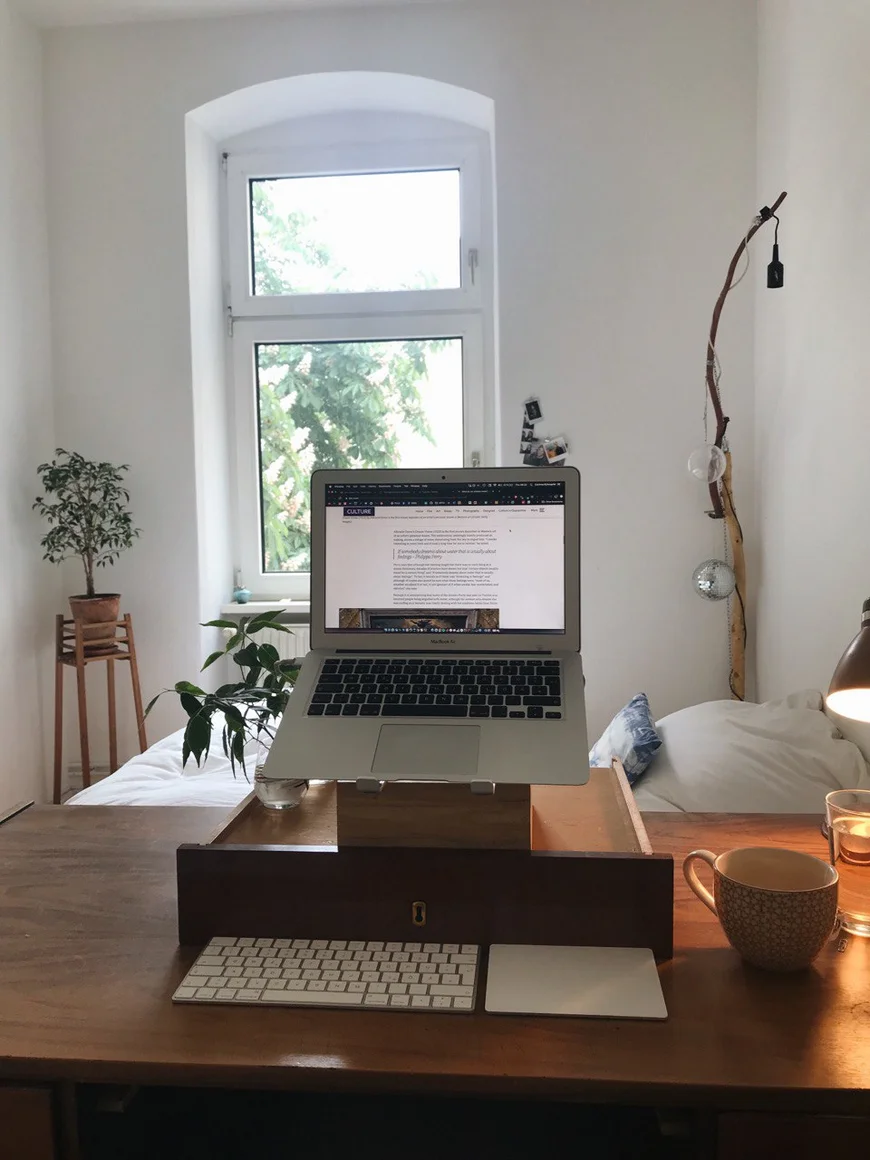 Schreibtisch mit einer umgedrehten alten SChublade darauf, die als Laptopständer dient. Dahinter sieht man ein bett und ein Fenster