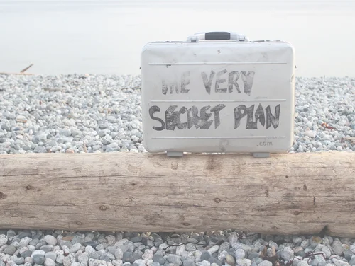 Im Hintergrund ist ein Kieselstrand und Wasser erkennbar. Im Vordergrund liegt ein Baumstamm auf dem steht ein grauer Metall·koffer mit dem Schriftzug The very secret plan