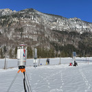 Field site Graswang: Laser scanner on a tripod standing on a flat valley bottom with snow and forested mountains in the background. Several scientists are working with different equipment in the valley. | Photo: Cosmic Sense Consortium