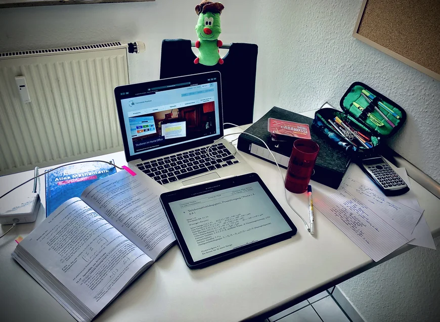 Auf einem Schreibtisch sieht man von oben einen Laptop, ein Tablet, Bücher, eines ist aufgeschlagen, es sind mathematische Formeln zu sehen, eine Fedrmappe und ein Ordner sowie Notizzettel und Taschenrechner