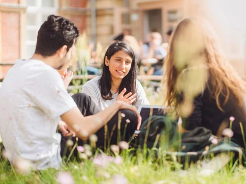 Three students sitting on the lawn on Am Neuen Palais campus talking to each other.