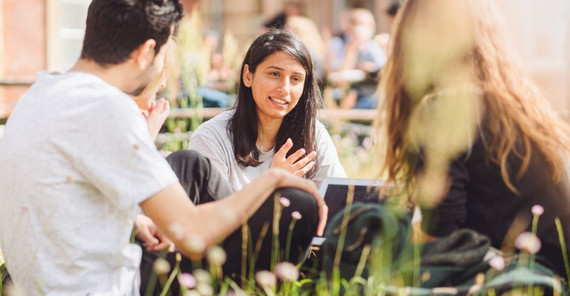 Drei Studierende sitzen auf dem Campus Neues Palais auf einer Wiese und unterhalten sich.