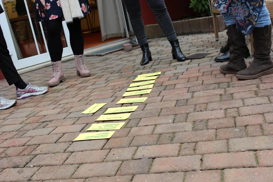 Das Bild zeigt einen Workshop. Eine Gruppe von Personen steht im Kreis auf einem gepflasterten Boden. Im Mittelpunkt liegen mehrere gelbe Karten mit grüner Schrift, die thematisch sortiert sind.