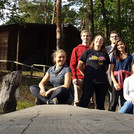 Zu sehen ein Gruppenbild des Jazzchors Campus Golm bei der Chorfahrt im Sommer 2021. Die 13 Teilnehmer*innen der Chorfahrt sitzen auf Holzbänken der Jugendherberge, die idyllisch im Wald gelegen ist.