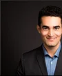 Portraitfoto eines Mannes mit schwarzen, kurzen Haaren, bis zur Schulter abgebildet. Er trägt eine schwarzes sakko und darunter ein weißes Hemd. Der Hintergrund ist schwarz.