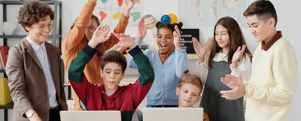 Schülerinnen und Schüler sowie eine Lehrkraft schauen zwei Schülern über die Schulter. Beide sitzen vor Laptops, einer freut sich zusammen mit den Umstehenden.