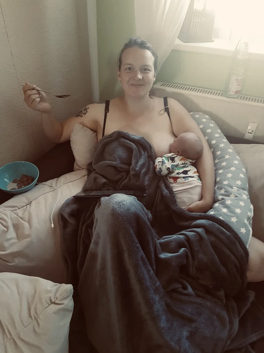 in einer Sofaecke sitzt eine junge Frau, sie hat ein Tablet auf dem Schoß und stillt gleichzeitig ihr Baby
