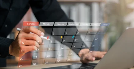 Mensch steht vor einem Tisch, wo ein Laptop von hinten zu sehen ist. Davor ist ein digitaler Kalender zu sehen.