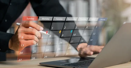 Mensch steht vor einem Tisch, wo ein Laptop von hinten zu sehen ist. Davor ist ein digitaler Kalender zu sehen.