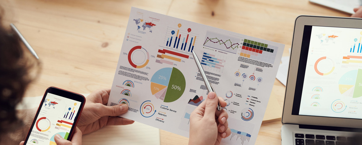 Über die Schultern zweier Menschen sieht man, wie sie sich mit den vor ihnen liegenden statistischen Graphen beschäftigen. - 