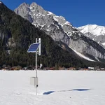 Field site Leutasch: CRNS sensor with solar panels installed at a flat valley bottom with snow. In the background are houses and forrested mountains | Photo: Cosmic Sense Consortium
