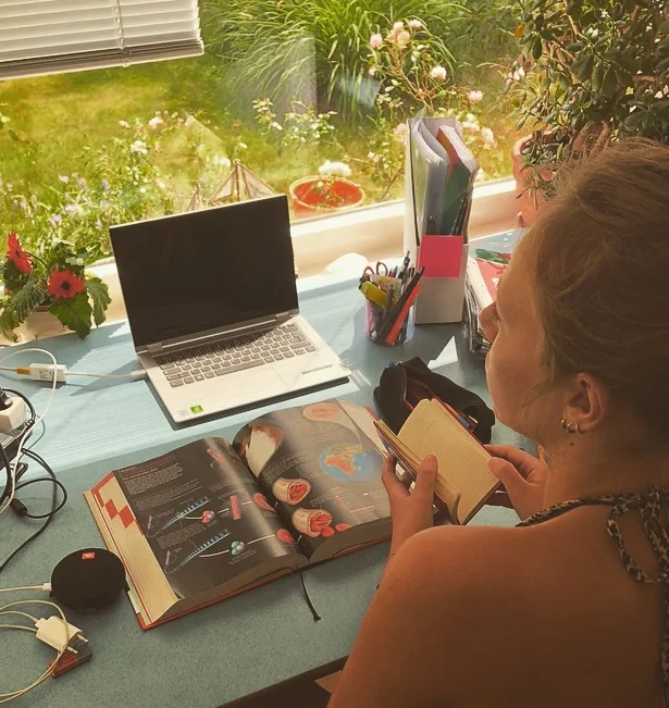 Mensch vor Schreibtisch. Darauf Buch, Laptop. Blick durch Fenster in Garten