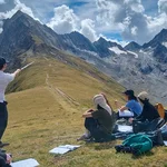 People sitting at a mountain ridge, one person is standing in front of them, explaining while gesturing.
