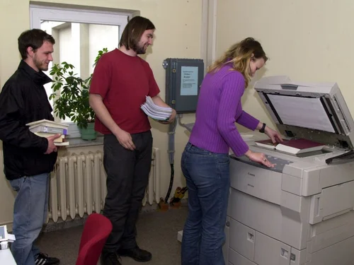 Das Bild zeigt Personen an einem Drucker. Eine Frau macht eine Kopie von einem Buch. Sie druckt eine Seite aus.