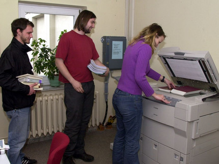 Das Bild zeigt Personen an einem Drucker. Eine Frau macht eine Kopie von einem Buch. Sie druckt eine Seite aus.